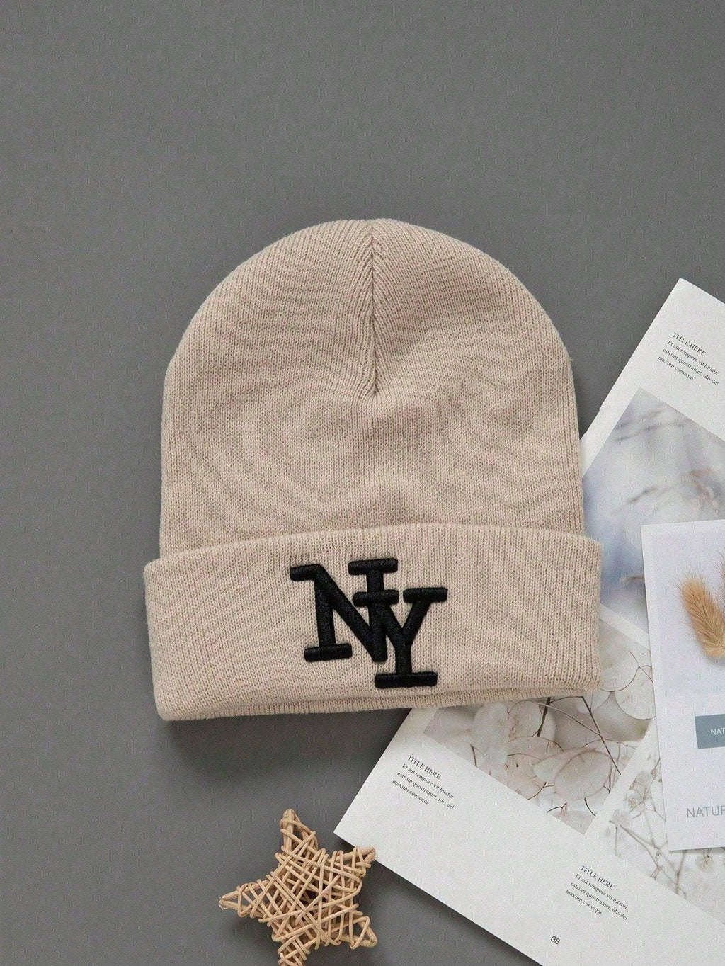 Beige cotton NY beanie with black embroidery, flat lay on gray background with magazine pages